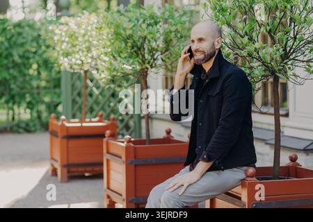 Joyeux homme d'âge moyen parlant sur smartphone à l'extérieur assis près de jardinières dans Sunny Garden Courtyard avec espace de copie gauche Banque D'Images