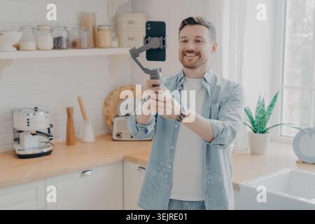 Jeune homme joyeux filmant avec smartphone sur cardan portable dans la cuisine lumineuse de la maison créant du contenu vlog de médias sociaux Banque D'Images