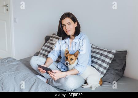 Femme Millennial détendue utilisant une tablette sur le lit à la maison avec petit chien reposant à côté d'elle dans l'intérieur confortable de la chambre lumineuse Banque D'Images