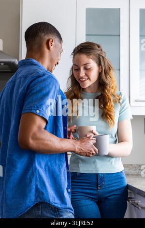 Couple diversifié échangeant des tasses en céramique côtelées tout en se tenant debout au comptoir de cuisine près de la cuisinière. Tasse, duo, armoire, verre, pierre, Banque D'Images