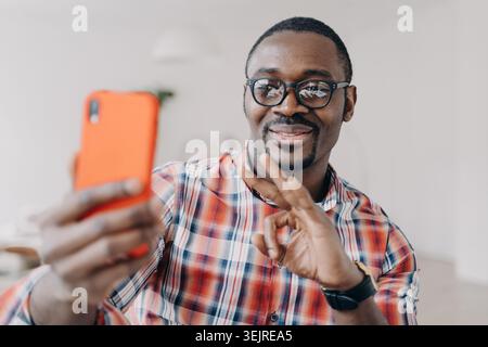 Joyeux jeune homme noir prenant smartphone Selfie à la maison tout en faisant Okay geste de main pendant le chat vidéo en ligne amical Banque D'Images