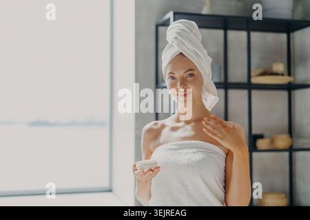 Sourire jeune femme appliquant la crème pour le visage dans la salle de bain lumineuse portant une serviette blanche après la douche pendant les soins de la peau frais du matin Routin Banque D'Images