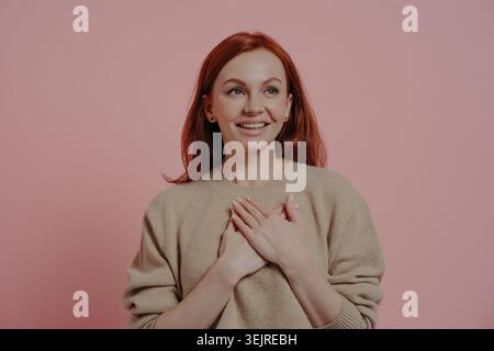 Femme millénaire aux cheveux rouges heureuse souriant avec les mains sur la poitrine contre fond rose montrant gratitude et émotion chaleureuse Banque D'Images