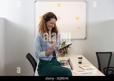 Femme assise sur le bord de la table de réunion, à l'aide de l'écran tactile avec stylet au bureau, portant une veste en denim. Professionnel, entreprise, mod Banque D'Images