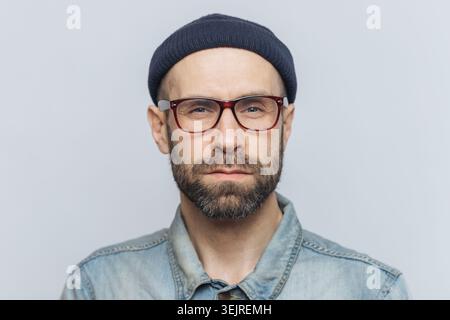 Homme d'âge moyen avec des lunettes et un bonnet regarde la caméra sur fond gris de studio Banque D'Images