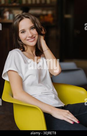Jeune femme dans un t-shirt blanc sourit en étant assise sur une chaise jaune à l'intérieur Banque D'Images