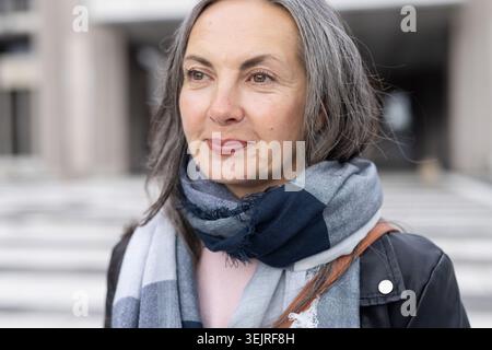 Femme adulte mature debout aux marches du bâtiment portant une écharpe rayée, une veste sombre et une sangle de sac. Portrait, senior, extérieur, urbain Banque D'Images