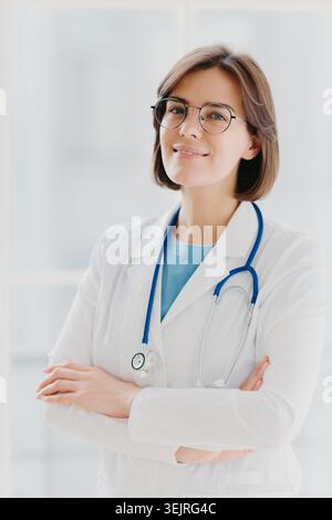 Femme médecin portant des lunettes et stéthoscope debout avec les bras croisés dans le bureau lumineux de la clinique Banque D'Images