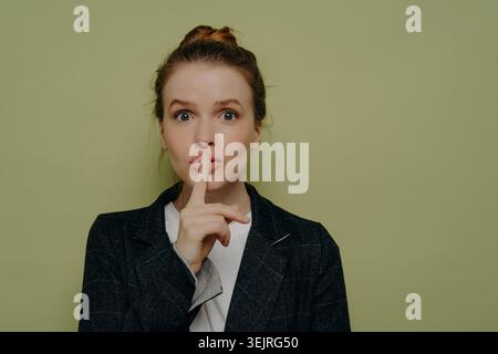 Jeune femme d'affaires avec le doigt aux lèvres faisant shh geste de silence avec de larges yeux regardant la caméra contre le fond vert sage Banque D'Images