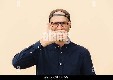 Homme d'âge moyen portant des lunettes couvrant la bouche avec la main contre le mur beige Banque D'Images
