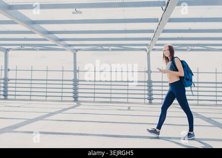 Jeune femme avec sac à dos marche à travers le pont urbain lumineux tenant smartphone Banque D'Images