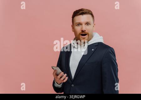 Surpris jeune homme tient smartphone tout en portant blazer et sweat à capuche contre le mur rose du studio Banque D'Images