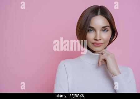 Jeune femme en col roulé blanc pose avec la main sur le menton contre le mur rose du studio Banque D'Images
