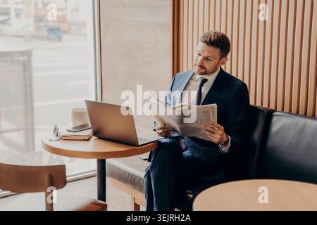 Homme d'affaires en costume bleu lisant le journal à côté de l'ordinateur portable dans l'espace de travail de café Banque D'Images