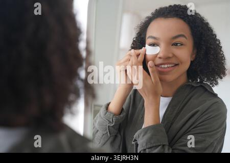 Jeune femme noire appliquant sous le cache-oeil tout en regardant dans le miroir de salle de bain Banque D'Images