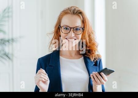 Souriante femme d'affaires aux cheveux rouges dans des lunettes tient smartphone dans un bureau lumineux Banque D'Images