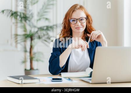 Femme d'affaires aux cheveux roux sourit tout en travaillant sur ordinateur portable au bureau dans le bureau lumineux Banque D'Images