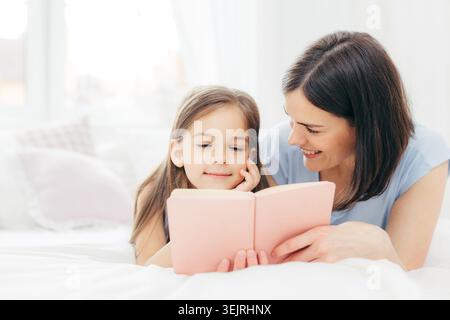 Mère et fille lisent un livre rose ensemble tout en étant allongées sur le lit à la maison Banque D'Images