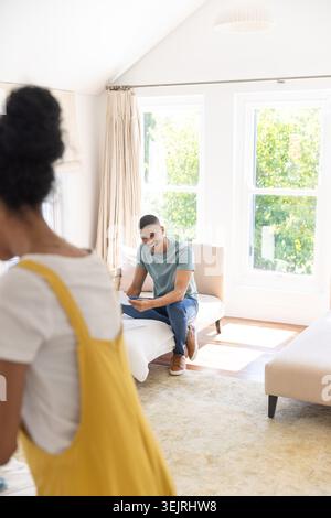 Homme de couple assis sur un canapé bas tenant des papiers, femme portant une robe jaune se déplaçant près de grandes fenêtres. Lit de jour, lumière du jour, ensoleillé, va Banque D'Images