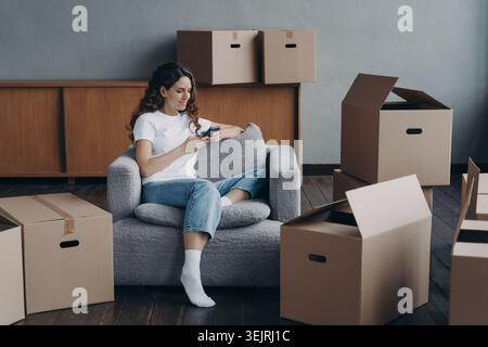 Jeune femme utilisant un smartphone tout en étant assise dans un fauteuil parmi les boîtes de déménagement dans le nouvel appartement Banque D'Images