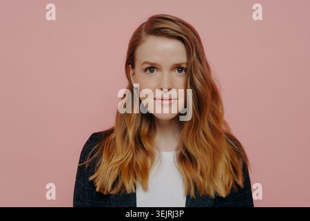 Jeune femme aux cheveux rouges en blazer foncé regardant directement la caméra avec une expression sérieuse sur fond de studio rose Banque D'Images