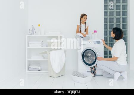 Mère et fille faisant la lessive ensemble à côté de la machine à laver dans une buanderie blanche lumineuse Banque D'Images