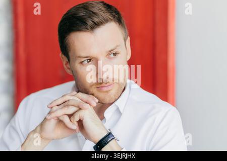 Homme d'affaires réfléchi en chemise blanche repose les mains tout en regardant loin près du mur rouge à l'intérieur Banque D'Images