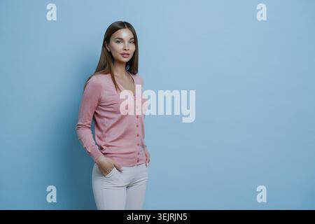 Jeune femme brune en cardigan rose se tient avec les mains dans les poches sur fond de studio bleu Banque D'Images