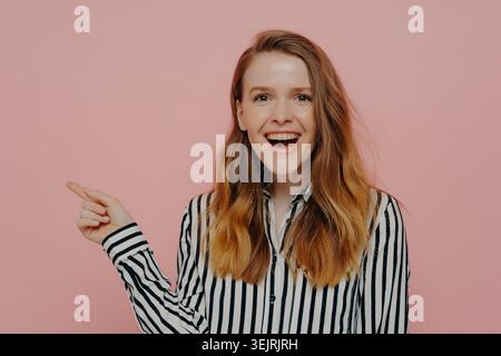 Jeune femme rousse pointe vers un espace de copie vide avec un sourire excité contre le mur rose du studio Banque D'Images