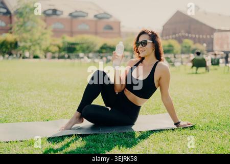 Femme en sportswear noir repose sur le tapis de yoga avec bouteille d'eau dans le parc ensoleillé de la ville Banque D'Images