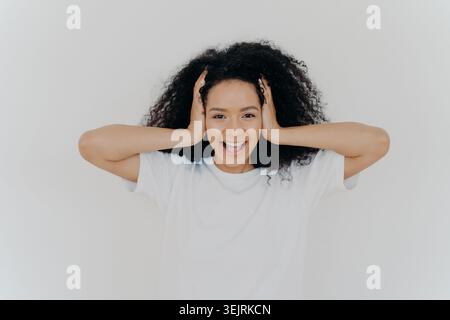 Jeune femme aux cheveux bouclés sourit tout en tenant sa tête contre le mur blanc du studio Banque D'Images