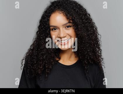 Jeune femme souriante avec de longues boucles naturelles pose en chemise noire contre le mur gris du studio Banque D'Images