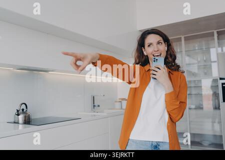 Jeune femme tenant smartphone et pointant dans la cuisine blanche moderne Banque D'Images