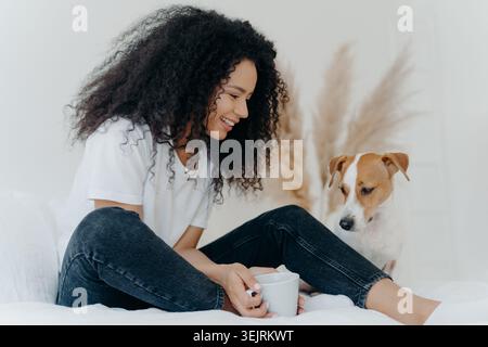 Jeune femme souriante assise sur le lit avec un Jack Russell Terrier tout en tenant une tasse à la maison Banque D'Images