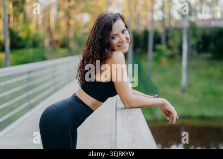 Jeune femme souriante en sportswear noir repose sur la balustrade de pont dans le parc vert Banque D'Images