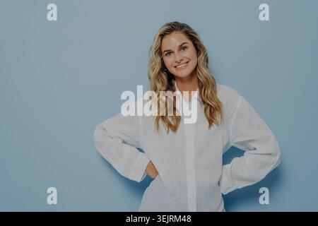 Jeune femme blonde en chemise blanche oversize souriant avec confiance avec les mains sur les hanches sur fond bleu clair Banque D'Images