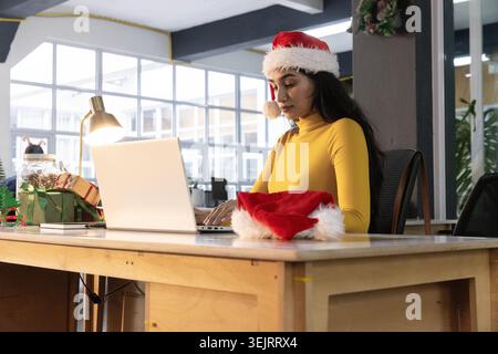 Femme indienne tapant sur un ordinateur portable argenté au bureau ouvert dans un pull moutarde, chapeau de Père Noël, espace de copie Banque D'Images