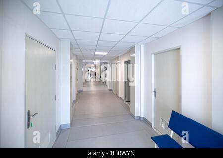 Un couloir d'hôpital intérieur long et lumineux avec des murs peints en blanc, des portes en bois fermées, un plafond suspendu avec des lumières carrées, un banc bleu sur la R Banque D'Images