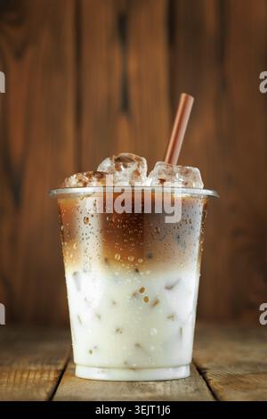 boisson au café latte glacé dans une tasse en plastique transparent avec de la paille brune, des glaçons et de la condensation, en restant sur fond de bois Banque D'Images