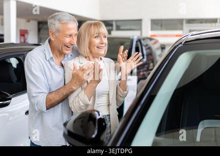 Couple d'aînés excités réagissant avec surprise en regardant la voiture dans la salle d'exposition de l'établissement concessionnaire Banque D'Images
