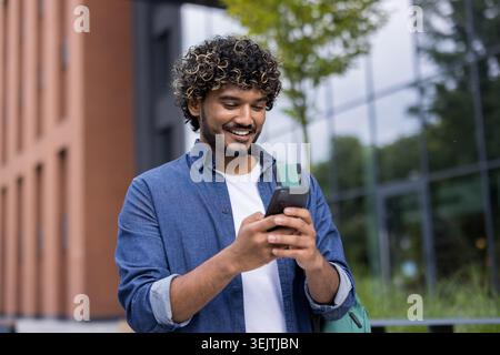 Jeune homme aux cheveux bouclés souriants, debout à l'extérieur près d'un bâtiment moderne tout en utilisant activement un smartphone, représentant la connexion, la communication décontractée et la technologie mobile contemporaine Banque D'Images