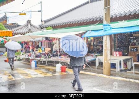 Un jour pluvieux sur un marché coréen traditionnel est capturé ici. Les gens avec des parapluies passent devant des stands colorés, avec des gouttes de pluie visibles ajoutant de l'humeur. Banque D'Images