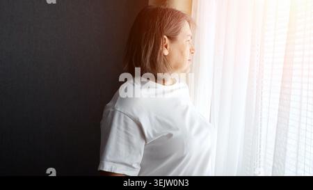 Femme asiatique mature debout près d'une fenêtre, regardant dehors avec une expression sombre, reflétant des sentiments d'isolement, de tristesse et de dépression en douceur Banque D'Images