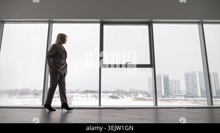 Femme d'affaires confiante marchant vers l'avant dans un espace de bureau moderne, contemplant le paysage urbain avec de hauts bâtiments et un paysage hivernal enneigé Banque D'Images
