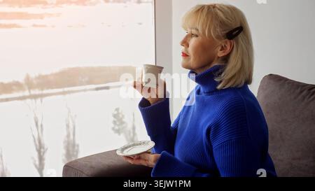 Femme senior dans un pull bleu est assis sur un canapé sirotant un café, regardant à travers une fenêtre un paysage enneigé, profitant d'un moment paisible et réfléchissant Banque D'Images