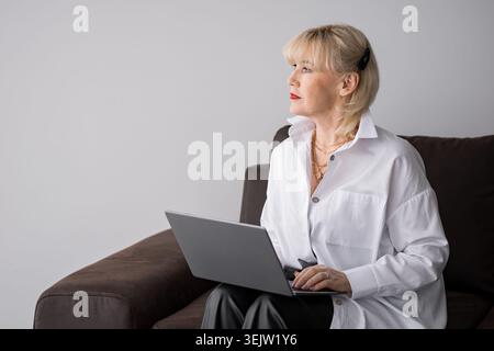 Femme d'affaires mature avec les cheveux blonds et rouge à lèvres assis sur un canapé, vue de côté, travaillant sur un ordinateur portable et regardant soigneusement loin tout en planifiant Banque D'Images