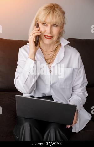 Femme âgée assise sur un canapé, tenant un ordinateur portable et s'engageant dans une conversation téléphonique, dépeignant le travail à distance efficace et les affaires modernes Banque D'Images