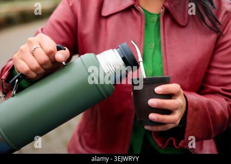 Femme dans une veste en cuir rouge versant de l'eau chaude d'un thermos vert dans une gourde traditionnelle mate Banque D'Images
