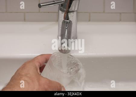 L'eau coule régulièrement du robinet chromé. L'évier blanc et le mur carrelé forment une toile de fond propre. Aucune émotion montrée, juste une action pratique. Idéal pour les routi quotidiens Banque D'Images
