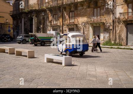 Deux femmes traversent une place historique ensoleillée en Italie, passant devant un taxi bleu à trois roues Piaggio qui passe devant un camion de déchets au milieu de T. Banque D'Images